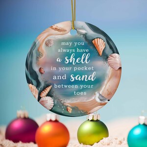 May include: A round, white ceramic ornament with a blue and brown seashell design. The ornament has the text "may you always have a shell in your pocket and sand between your toes".
