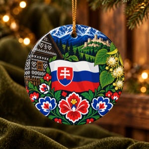 Op de afbeelding: Een ronde keramische hanger met een kleurrijke illustratie van de Slowaakse vlag boven een folkloristisch bloemenpatroon. Het ontwerp bevat een traditioneel houten huis, een stenen kasteel op een heuvel, blauwe bergen en lindenbloesems.
