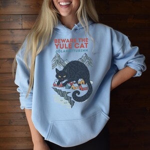 May include: A light blue hooded sweatshirt featuring a graphic print of a large black cat looming over a small village of colorful houses nestled beneath mountains. The text reads BEWARE THE YULE CAT and J&Oacute;LAK&Ouml;TTURINN in red and blue lettering.