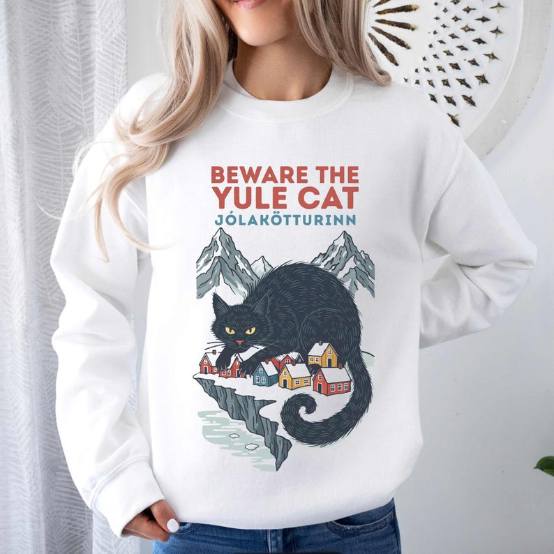 May include: A white crewneck sweatshirt featuring a graphic of a large black cat with yellow eyes looming over a small village of colorful houses on a snowy cliff, with mountains in the background and the text BEWARE THE YULE CAT J&Oacute;LAK&Ouml;TTURINN.