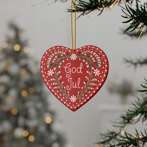 May include: Red heart-shaped Christmas ornament with the words "God Jul" in white script. The ornament features a white dotted border and is decorated with white flowers and green leaves. It hangs from a gold string.