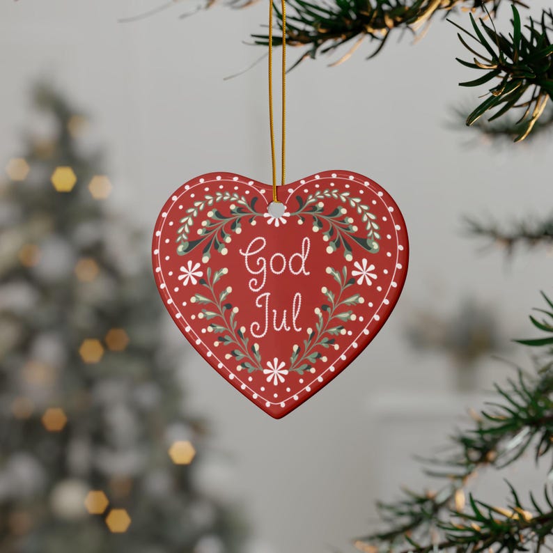 May include: Red heart-shaped Christmas ornament with the words "God Jul" in white script. The ornament features a white dotted border and is decorated with white flowers and green leaves. It hangs from a gold string.