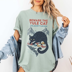 May include: A sage green short-sleeve t-shirt featuring a graphic of a large black cat looming over a small village with mountains in the background. The text reads BEWARE THE YULE CAT J&Oacute;LAK&Ouml;TTURINN in red and blue lettering on the front.
