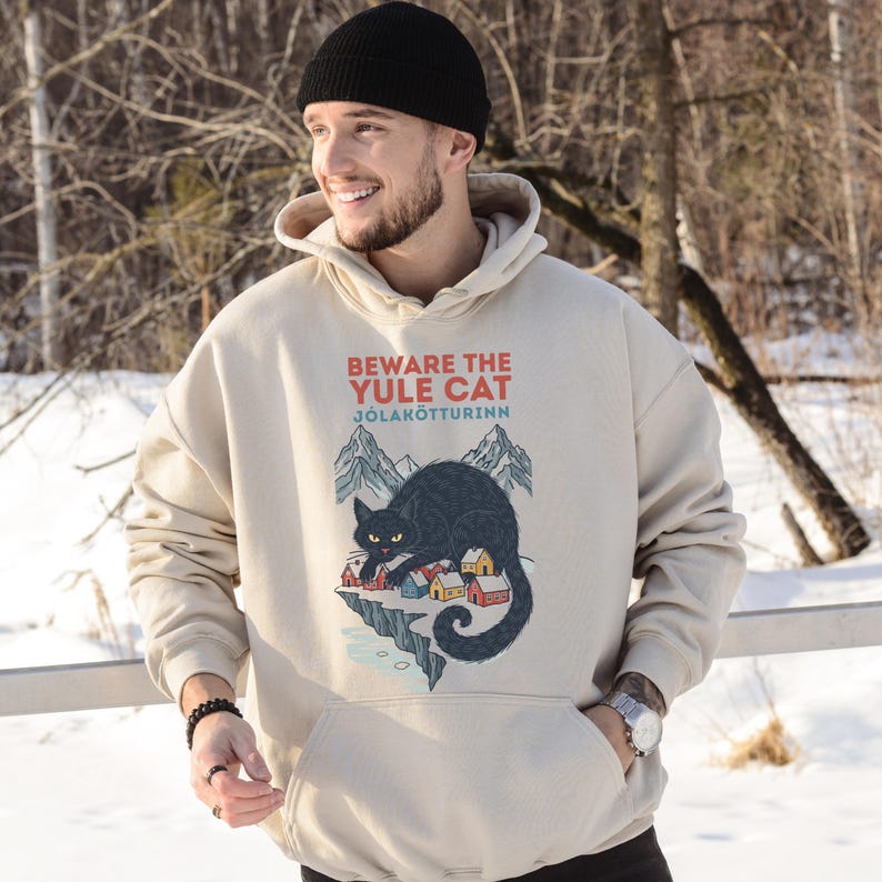 May include: A beige hooded sweatshirt featuring a graphic print of a large black cat looming over a small village with mountains in the background. The text reads BEWARE THE YULE CAT and J&Oacute;LAK&Ouml;TTURINN in red and blue lettering above the illustration.