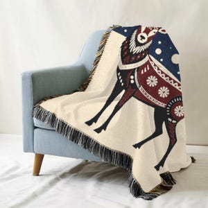 Puede incluir: Una manta tejida con fondo crema y un dise&ntilde;o de reno estilizado en negro y rojo. El reno tiene un cuerpo con estampado rojo y blanco y una cabeza con estampado azul y blanco. La manta tiene flecos en todos los lados.