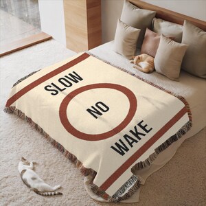 May include: A cream-colored woven throw blanket with a red stripe and a circle with the text "NO WAKE" inside. The text "SLOW" is above the circle.