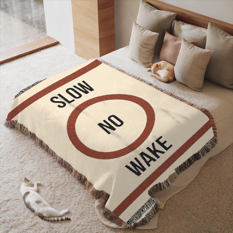 May include: A cream-colored woven throw blanket with a red stripe and a circle with the text "NO WAKE" inside. The text "SLOW" is above the circle.