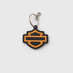 May include: Black and orange Harley-Davidson logo keychain. The keychain features a shield-shaped emblem with a horizontal bar, a classic design. The keychain is attached to a silver metal ring.