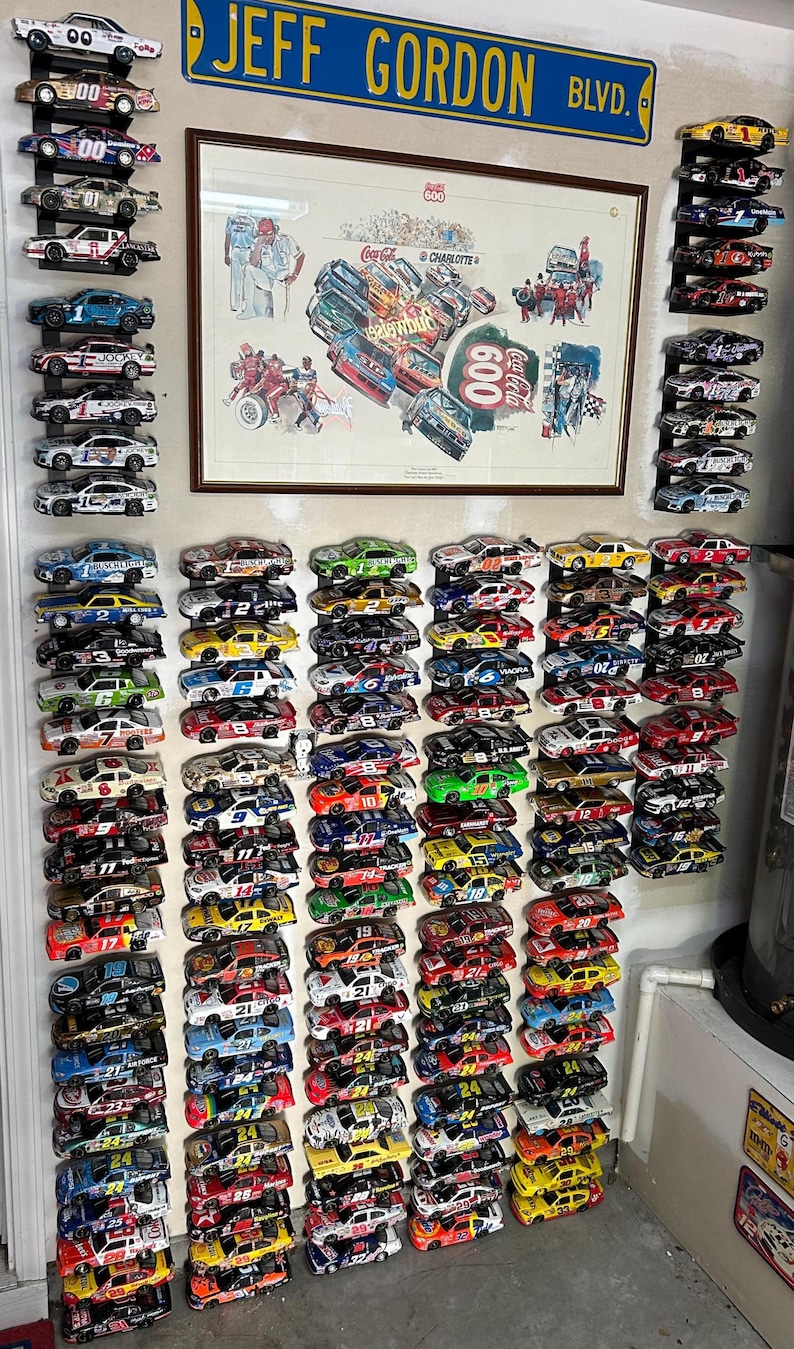 May include: A collection of miniature race cars, many with sponsor logos and racing numbers, displayed on a wall. A framed print and a street sign that reads "JEFF GORDON BLVD." are also visible. The cars are arranged in rows.