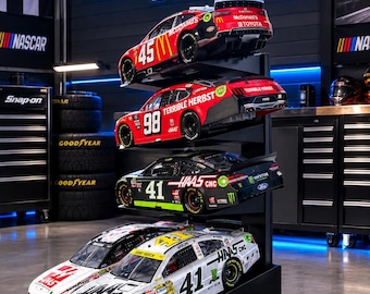 May include: A black metal display stand holding five diecast stock car models. The top three cars are stacked vertically, while two cars sit side-by-side at the base. The stand features the text DIECAST STANDS.COM in blue neon lettering above the cars.
