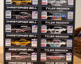 May include: A black vertical display rack containing ten NASCAR die-cast race cars in individual boxes. The left column features Christopher Bell's cars, and the right column features Tyler Reddick's cars, each labeled with their name and year.