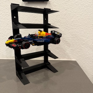 May include: Black metal display stand with three shelves holding a blue, red, and yellow race car. The stand is mounted on a white wall with a black bracket above it. The race car has sponsor logos.