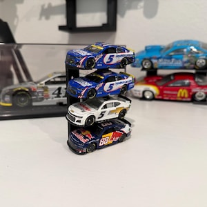 May include: A collection of miniature race cars in various colors, including blue, red, white, and black. Some cars are stacked on a black display stand, while others are displayed in a clear case. The cars feature sponsor logos and racing numbers.