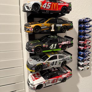 May include: A collection of NASCAR race cars displayed on black shelves. The cars feature various sponsor logos and racing numbers, including a red and white car with the number 45 and a McDonald's logo.