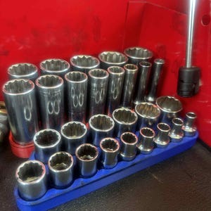 May include: A collection of chrome sockets of various sizes, neatly arranged in a blue holder. The sockets are cylindrical with a star-shaped opening. A red toolbox is in the background.