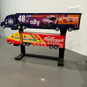 May include: Two toy semi-trucks on a black stand. The top truck is dark blue with "48 ally" and "ALEX BOWMAN" text. The bottom truck is red and yellow with "Kellogg's Racing" text.