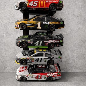 May include: A display of five NASCAR race cars on a black shelf against a gray wall. The cars feature sponsor logos like McDonald's, Haas CNC, and Gearwrench, with prominent numbers such as 45 and 41. Each car has detailed paint schemes.