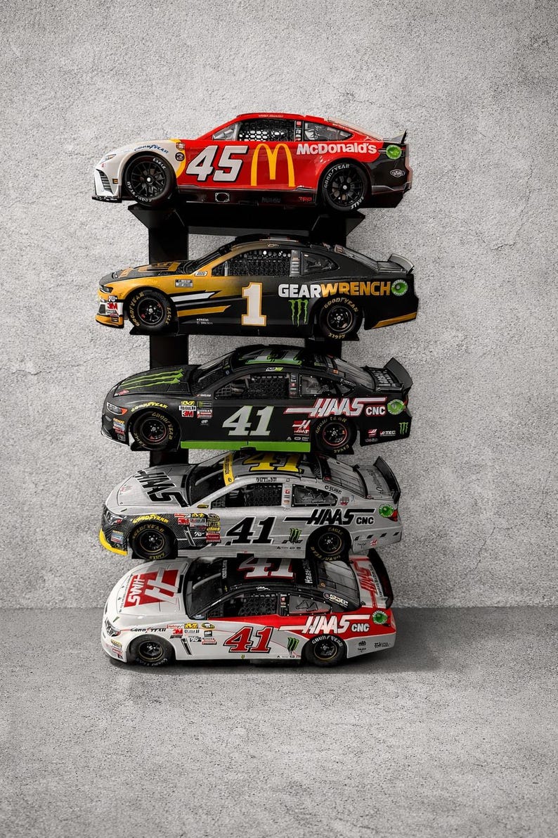 May include: A display of five NASCAR race cars on a black shelf against a gray wall. The cars feature sponsor logos like McDonald's, Haas CNC, and Gearwrench, with prominent numbers such as 45 and 41. Each car has detailed paint schemes.