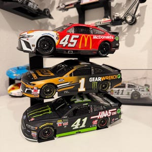May include: Three NASCAR model cars stacked on a shelf. The top car is red and white with the number 45 and McDonald's branding. The middle car is black and gold with the number 1. The bottom car is black with the number 41.