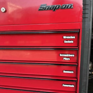 May include: A red Snap-on tool chest with black trim and drawer labels. The labels read "Wrenches, Sockets, Screwdrivers, Pliers, Hammers, and Powertools."