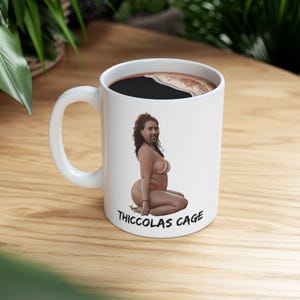 Op de afbeelding: Witte keramische koffiemok gevuld met donkere vloeistof. De mok heeft een humoristische illustratie van een persoon in een bikini met de tekst "Thiccolas Cage" onder de afbeelding. De mok heeft een wit handvat.