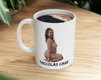 May include: White ceramic coffee mug filled with dark liquid. The mug features a humorous illustration of a person in a bikini with the text "Thiccolas Cage" printed below the image. The mug has a white handle.