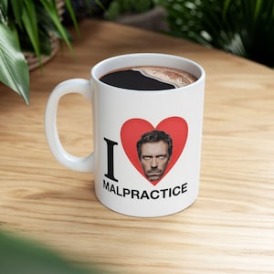 Puede incluir: Taza de café de cerámica blanca con interior negro llena de café. La taza presenta el texto "I MALPRACTICE" con un corazón rojo que contiene un retrato de un hombre.