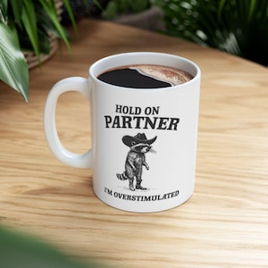 May include: White ceramic coffee mug with the text "HOLD ON PARTNER" and "I'M OVERSTIMULATED". The mug features a cartoon raccoon wearing a cowboy hat and boots. The mug is filled with dark coffee.