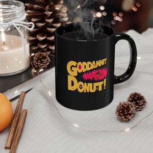 以下が含まれることがあります： 黄色とピンクで「GODDAMNIT DONUT！」の文字が入った黒いセラミックマグカップ。 マグカップには濃い液体が入っており、上から湯気が立っています。 マグカップは明るい灰色の表面にあります。