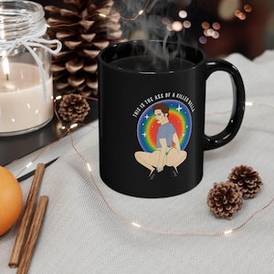 May include: Black ceramic mug featuring a cartoon illustration of a person squatting, set against a rainbow backdrop. The text "THIS IS THE ASS OF A KILLER BELLA" is printed around the image.
