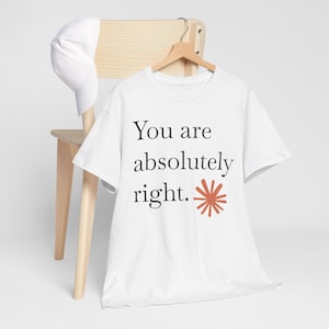 May include: A white short-sleeved t-shirt featuring the black text "You are absolutely right." and a small terracotta-colored starburst graphic. A plain white baseball cap is draped over the back of the wooden chair holding the shirt.