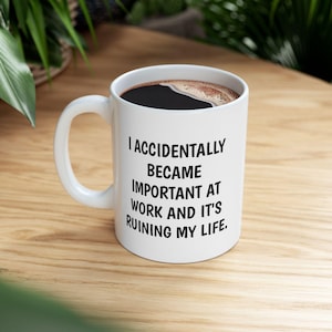 Pode incluir: Caneca de cerâmica branca cheia de café escuro. A caneca tem uma alça curva e apresenta o texto: "I ACCIDENTALLY BECAME IMPORTANT AT WORK AND IT'S RUINING MY LIFE." A caneca está em uma superfície de madeira.