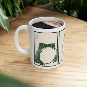 May include: White ceramic coffee mug featuring a green frog illustration in a Japanese art style. The mug has a black rim and is filled with dark coffee. The frog design is on a cream-colored background with a black border.