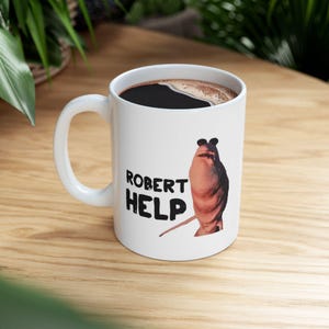 Puede incluir: Taza de cerámica blanca llena de café oscuro. La taza presenta una imagen de dibujos animados de una criatura parecida a una babosa y las palabras "ROBERT HELP" en texto negro. La taza tiene un asa blanca y está sobre una superficie de madera.