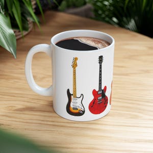May include: White ceramic coffee mug featuring illustrations of three electric guitars. The guitars are in various colors, including a sunburst, red, and orange. The mug is filled with dark coffee and sits on a wooden surface.