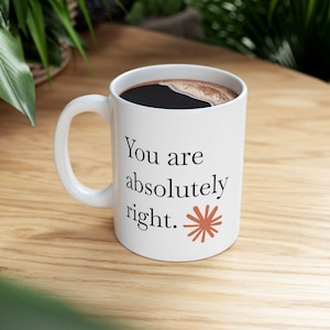 May include: A white ceramic mug filled with dark coffee. The exterior features the black printed text "You are absolutely right." followed by a small, terracotta-coloured starburst graphic. The mug has a curved handle on the left side.