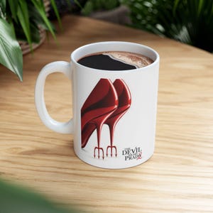 May include: White ceramic coffee mug with a handle, filled with dark coffee. The mug features a graphic of red high heels with trident-shaped heels, and the text "The Devil Wears Prada".