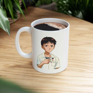 Op de afbeelding: Witte keramische mok met een anime-personage-illustratie. Het personage heeft donker haar en draagt een wit overhemd. De mok is gevuld met donkere koffie en heeft een wit handvat.