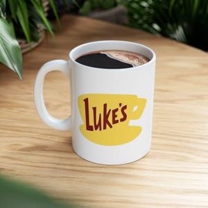 May include: White ceramic mug filled with dark coffee. The mug features a yellow coffee cup design with the text "Luke's" in a brown font. The mug has a white handle and sits on a wooden surface.