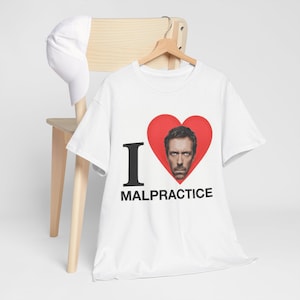 Puede incluir: Camiseta blanca con un corazón rojo que contiene un retrato y el texto "I MALPRACTICE". Una gorra de béisbol blanca cuelga de una silla de madera.