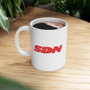 May include: White ceramic mug with the red letters "SDN" printed on the side. The mug is filled with dark coffee and sits on a wooden surface. The mug has a curved handle.