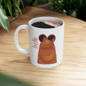 May include: White ceramic coffee mug with a cartoon illustration of a brown creature wearing sunglasses. The mug is filled with dark coffee and has the word "hello" printed on the side.