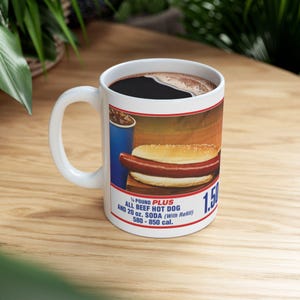 May include: White ceramic mug with a graphic of a hot dog, soda, and text. The text reads "¼ POUND PLUS ALL BEEF HOT DOG AND 20 oz. SODA (With Refill) 580-850 cal.". The mug is filled with dark liquid and has a white handle.
