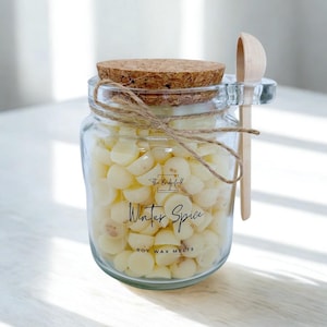 May include: A clear glass jar filled with small, pale yellow soy wax melts. The jar has a cork lid, a twine bow tied around the neck, and a small wooden spoon attached to the side. The label reads The Bridgefull Winter Spice SOY WAX MELTS.