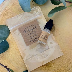 May include: A car diffuser refill bottle with a black cap and a small brown paper label. A clear plastic bag contains a brown paper card with the text "The Bridgefield Candle Company" and "Snow Pine Car Diffuser Refill".