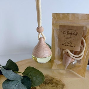 May include: A wooden car diffuser with a light brown wooden top and a clear glass bottom filled with a light yellow liquid. The diffuser is hanging from a white string and is packaged in a brown paper bag with the text "The Bridgefull" and "Spa Day Car Diffuser".