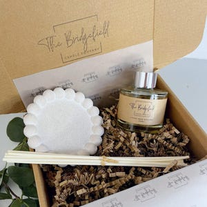 May include: A white ceramic diffuser dish, a clear glass bottle of diffuser oil with a silver cap, and a set of white diffuser sticks in a brown cardboard box with the text "The Bridgefield Candle Company" printed on the top. The box is filled with brown crinkled paper.