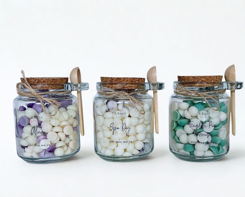May include: Three clear glass jars filled with small, hemispherical soy wax melts. Each jar has a cork lid, a small wooden spoon attached with twine, and a label reading Alien, Spa Day, or English Pear & Freesia. The wax melts are white, purple, and green.
