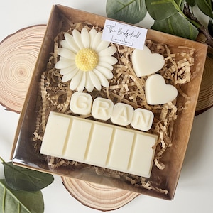 May include: A gift box filled with white wax melts, including a bar with the word "GRAN" spelled out, two heart shapes, and a white daisy. The box is lined with brown paper shreds.