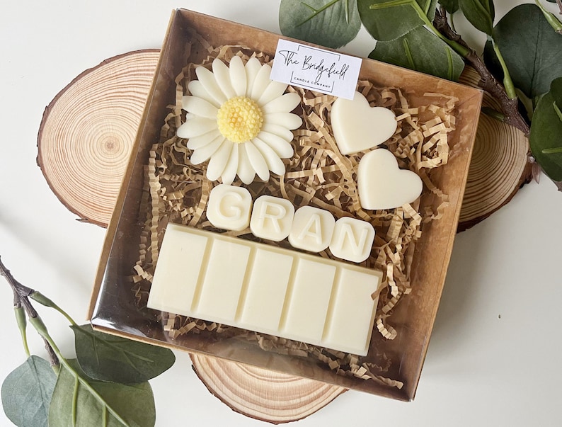 May include: A gift box filled with white wax melts, including a bar with the word "GRAN" spelled out, two heart shapes, and a white daisy. The box is lined with brown paper shreds.